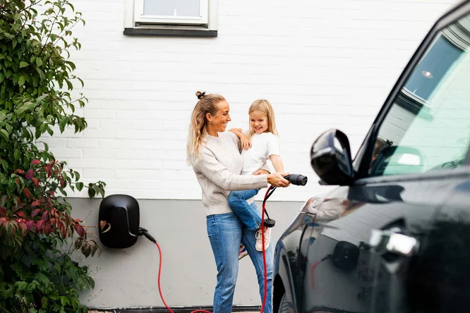 Imaginea arată o persoană care ține în brațe un copil în timp ce conectează un cablu de încărcare la un automobil electric. Scena are loc în exterior, lângă un perete alb din cărămidă, decorat cu vegetație verde și roșie în partea stângă. Deasupra celor doi se află o fereastră, iar stația de încărcare este montată pe perete, cu un cablu roșu care face legătura cu mașina.