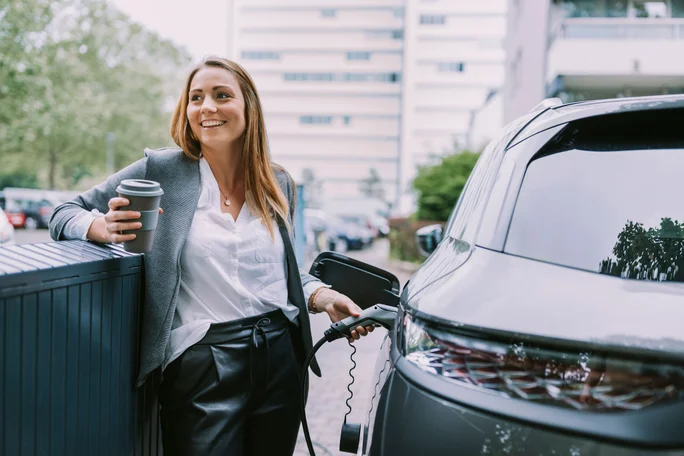Imaginea prezintă o persoană aflată lângă un vehicul electric aflat la încărcare. Persoana ține în mână o ceașcă de cafea și cablul de încărcare. În fundal se observă o clădire cu mai multe etaje, spații verzi și câteva mașini parcate. 