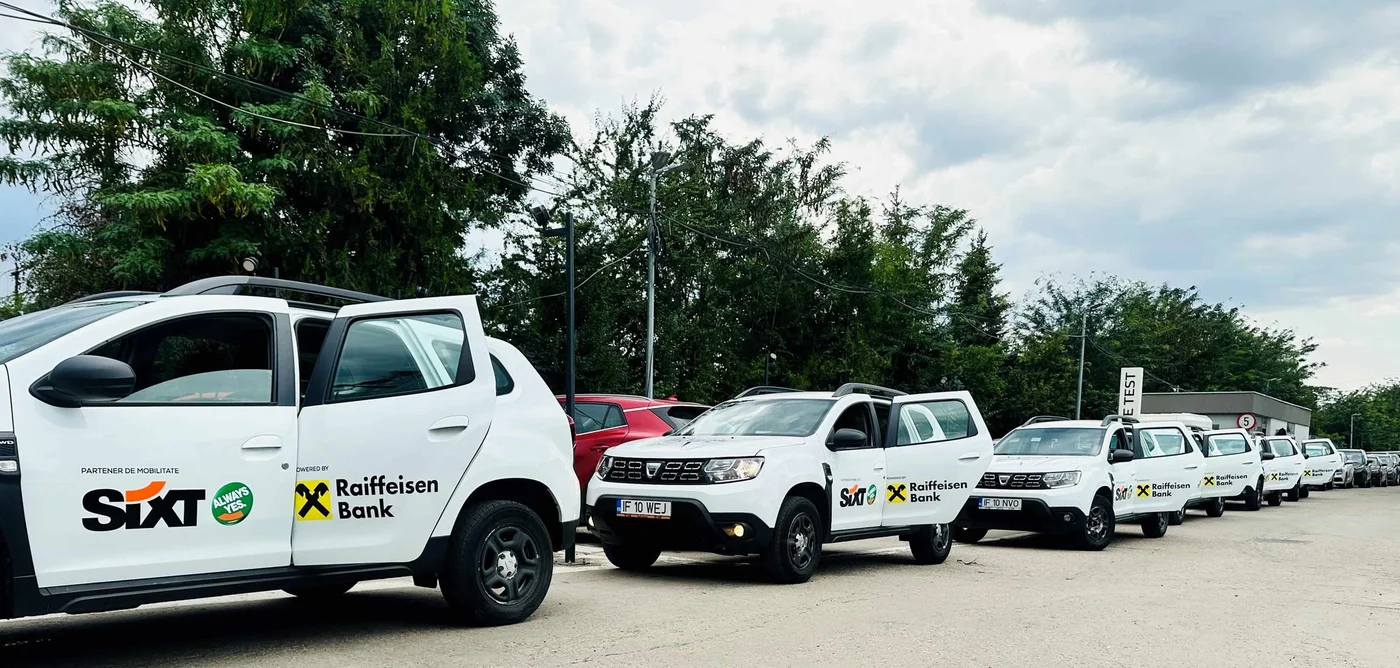 Imaginea prezintă un șir de SUV-uri albe parcate pe marginea unui drum. Vehiculele sunt branduite cu siglele „Sixt” și „Raiffeisen Bank”. În fundal se pot observa copaci, alte mașini și un cer înnorat.