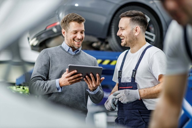 Imaginea prezintă două persoane într-un atelier sau garaj auto. Una dintre ele poartă un pulover gri peste o cămașă albastră deschisă și ține o tabletă, iar cealaltă este îmbrăcată în salopetă de lucru și poartă mănuși, sugerând implicarea în activități mecanice. În fundal se observă un automobil ridicat pe un elevator hidraulic.